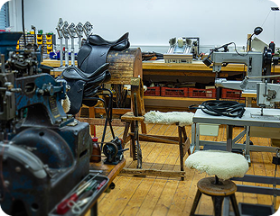 Werkstatt mit verschiedenen Werkzeugen und Maschinen zur Herstellung von Sätteln; im Vordergrund stehen eine Sitzgelegenheit und eine Nähmaschine, während im Hintergrund weitere Ausrüstungen sichtbar sind.